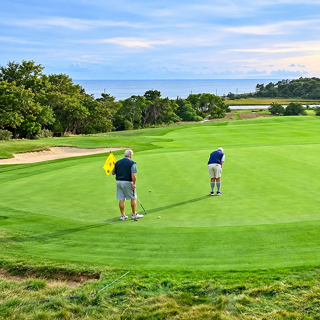 Two golfers putting on a green at a coastal golf course overlooking the ocean.