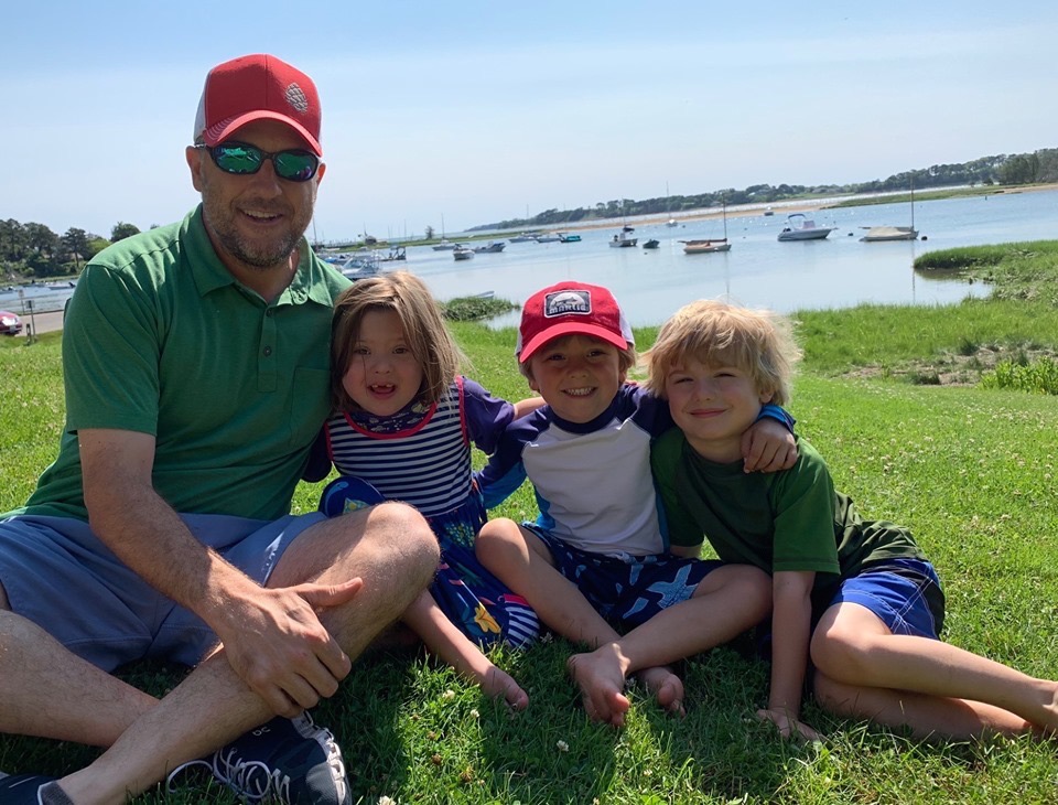 Dad_Kids a man with three children hugging and sitting down on the grass in front of water