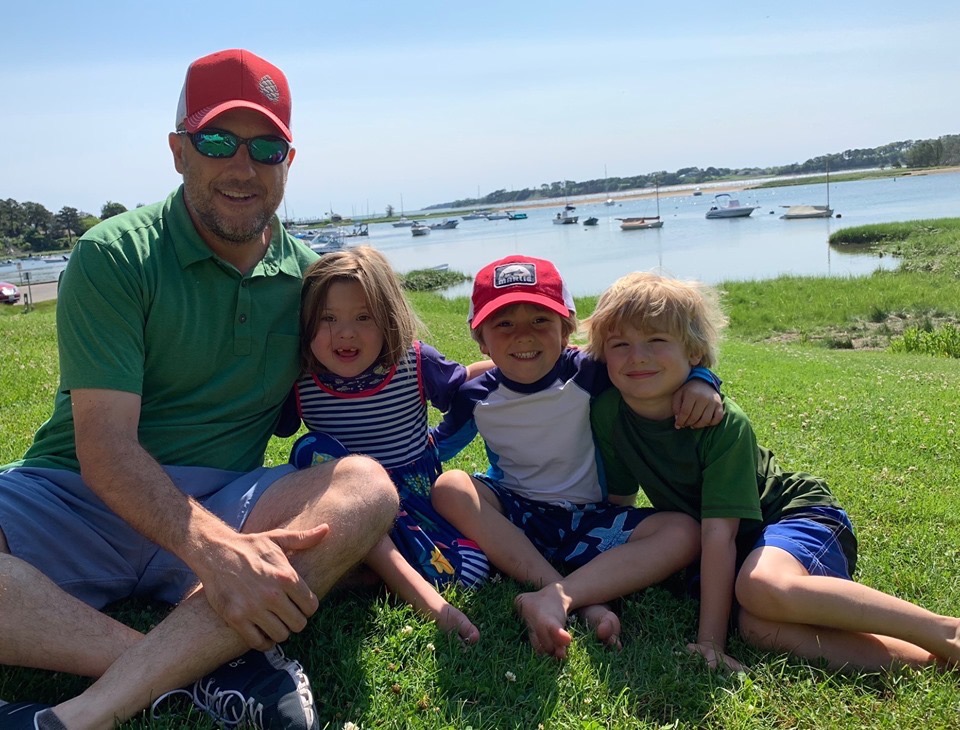 a man with three children hugging and sitting down on the grass in front of water