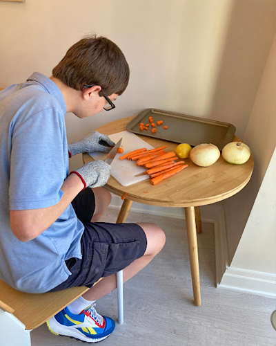 A male Best Buddie's Sleepover participant is cutting carrots and onions as he is being taught how to cook.