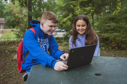 Young man in blue hoodie and young woman in purple hoodie smile at a computer while sitting at a table outside