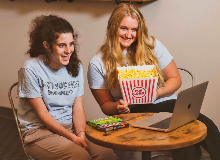 Two female Best Buddies participants sitting at a small table, where they are sharing popcorn while engaged virtually on a laptop computer.