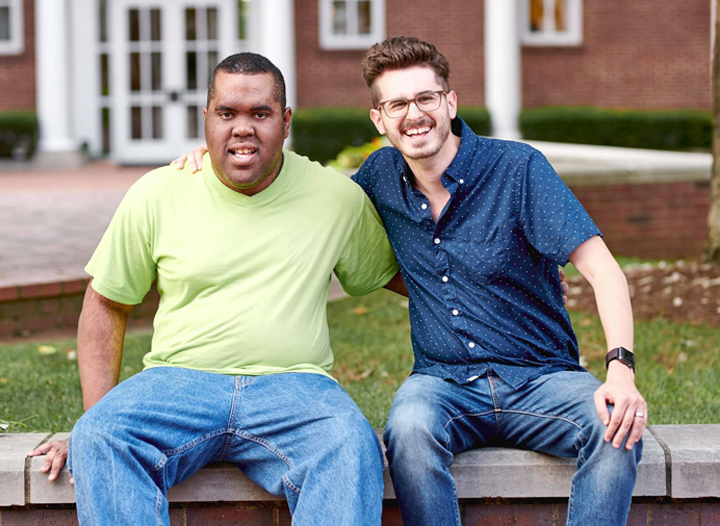 Two male Best Buddies participants pose with their arms around their shoulders in front of a brick building.