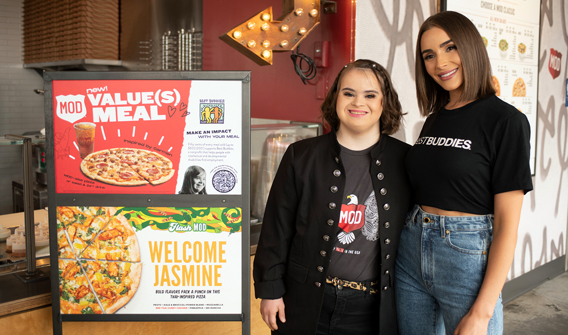 Best Buddies Supporter, Olivia Cumpo, and a female Best Buddies participant are shown inside a MOD's Pizza, next to a Mod's Pizza Value Meal sign.