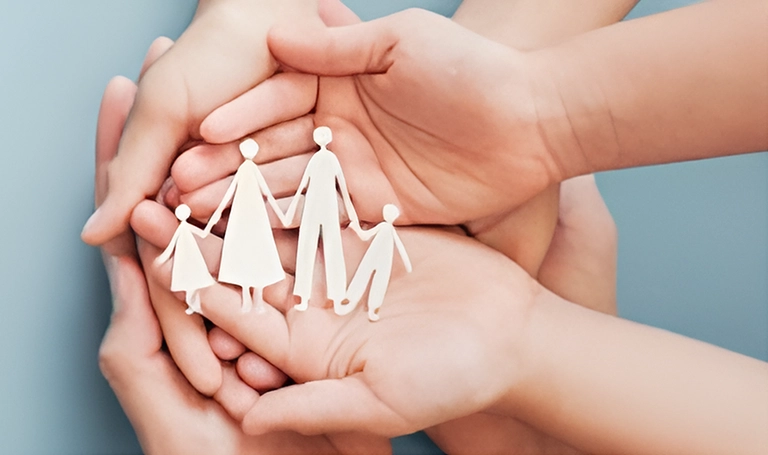 A circle of multiple hands are holding a paper cut-out of a family silhouette consisting of a nuclear family.