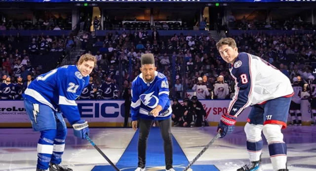 Best Buddies Ambassador poses with two Tampa Bay Lightning hockey players on the ice in front of a large crowd.