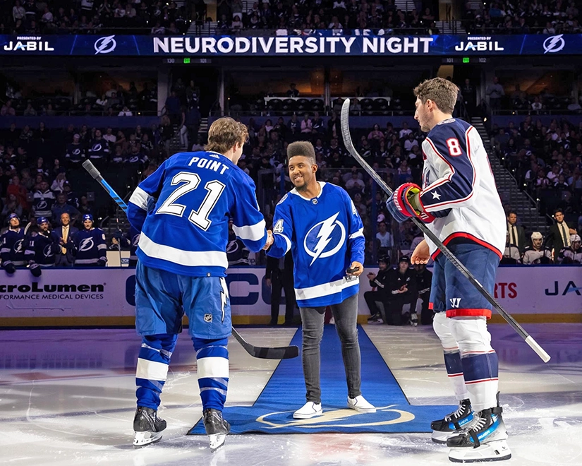 Joshua Felder, Best Buddies Ambassador, stands alongside two hockey players on the ice rink, exchanging handshakes with one of the players, captivating a large crowd.
