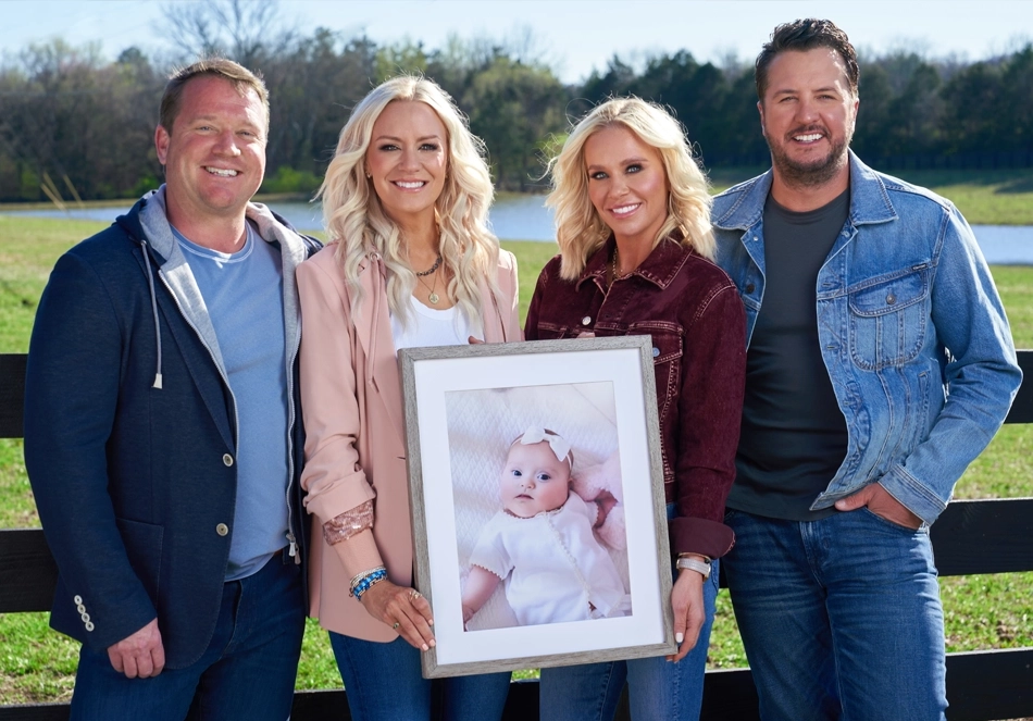 Outside alongside a wooded fence overlooking a pond and wooded area, there are 4 people posing for a picture. From left to right: Bo and his wife Ellen Boyer, who proudly holds a picture of their daughter Sadie Brett Boyer, standing alongside Caroline and Country music artist Luke Bryan.