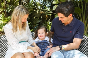 In a tropical outdoor setting, a mother and father is smiling, while holding their daughter with Down Syndrome. 