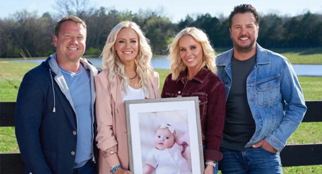 Outside alongside a wooded fence overlooking a pond and wooded area, there are 4 people posing for a picture. From left to right: Bo and his wife Ellen Boyer, who proudly holds a picture of their daughter Sadie Brett Boyer, standing alongside Caroline and Country music artist Luke Bryan.