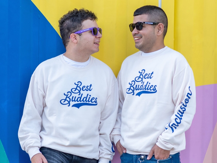 Two Best Buddies program participants looking at each other wearing sunglasses and sweatshirts that say Best Buddies.