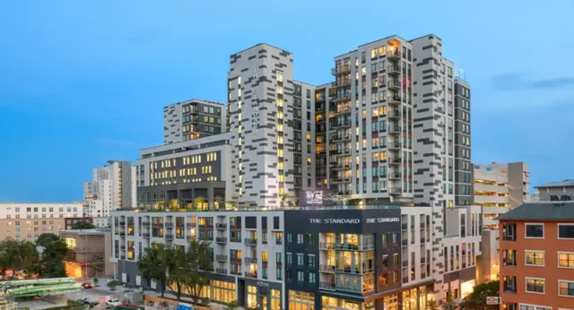 Modern high-rise apartment complex in Austin, Texas, featuring mixed facade design with glass and concrete, multiple balconies, and illuminated city skyline at dusk.
