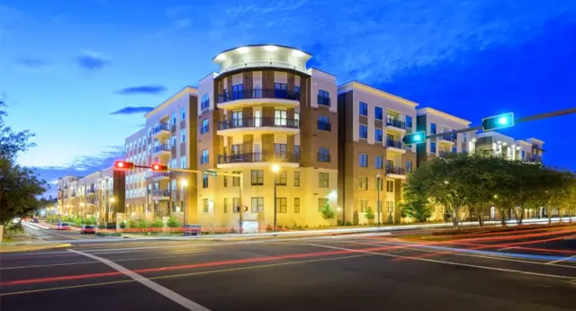 Exterior view of The Strand Tallahassee modern apartment building on a corner intersection at dusk with city lights.