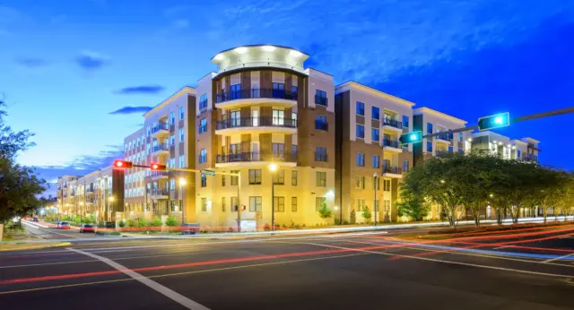 2026 BBI News Modern apartment building at a city intersection at dusk, with illuminated balconies, streetlights, and light trails from passing traffic.
