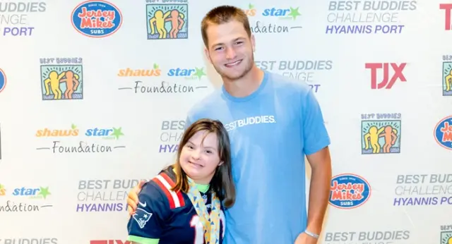 News - National Drake Maye meets with Best Buddies featured image The image shows a young man and a young girl smiling together in front of a step-and-repeat backdrop featuring Best Buddies Challenge: Hyannis Port branding along with sponsor logos like Jersey Mike’s, TJX, and Shaw’s Star Market Foundation. The man is wearing a light blue “Best Buddies” t-shirt and has his arm around the girl, who is wearing a sports jersey and several medals around her neck. Both appear happy and relaxed, highlighting a friendly and supportive moment at a Best Buddies event.