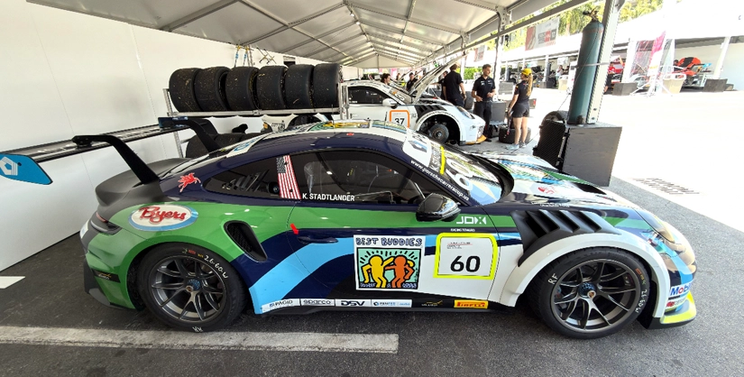 The image shows a side view of a professional race car parked inside a covered pit or garage area at a track. The car is a Porsche-style race car with the number 60 displayed on the door, along with prominent Best Buddies branding featuring the recognizable logo of two stylized figures. The car has a bold, multicolor livery in shades of green, blue, and white, with additional sponsor decals along the side. The name “K. Stadtlander” is printed on the side window, identifying the driver. In the background, several people are working around other race cars, and stacks of racing tires are visible, indicating an active race-day or practice environment.