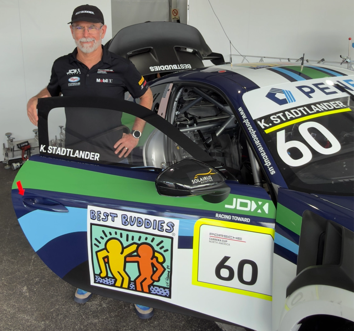 The image shows a man standing next to a race car with the driver-side door open. He’s wearing a black cap and a black polo shirt with sponsor logos, and he’s smiling slightly with one hand on his hip. The car appears to be a professional race car, featuring the number 60 and branding for Best Buddies on the door panel. The Best Buddies logo—two stylized figures in yellow and orange—is prominently displayed. Above the door, the name “K. Stadtlander” is printed, indicating the driver. A red arrow in the image points toward the car’s door handle area. The setting looks like a garage or pit area, with tools and equipment faintly visible in the background.