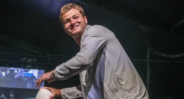 Drake Maye, smiling, stands on stage at a Best Buddies event while holding a football above a podium displaying the Best Buddies logo. Stage lighting, large screens, and event attendees are visible in the background, creating an energetic atmosphere as he appears to participate in an onstage activity.