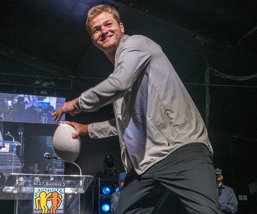 The 27th Annual Best Buddies Challenge: Hyannis Port Drake Maye, smiling, stands on stage at a Best Buddies event while holding a football above a podium displaying the Best Buddies logo. Stage lighting, large screens, and event attendees are visible in the background, creating an energetic atmosphere as he appears to participate in an onstage activity.