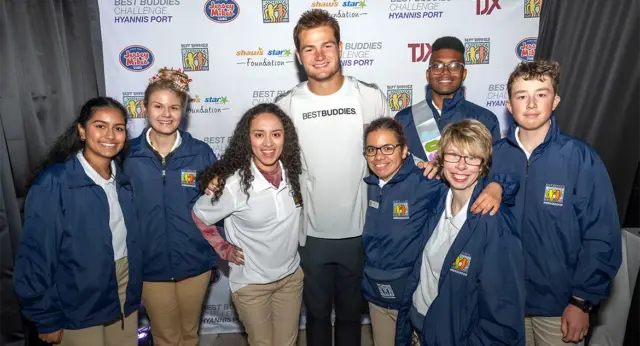 A group of Best Buddies Ambassadors pose with Drake Maye in front of a Best Buddies Challenge Hyannis Port step-and-repeat backdrop featuring sponsor logos. The group smiles for a photo, wearing matching ambassador jackets and event attire, while standing together at the annual fundraising event.