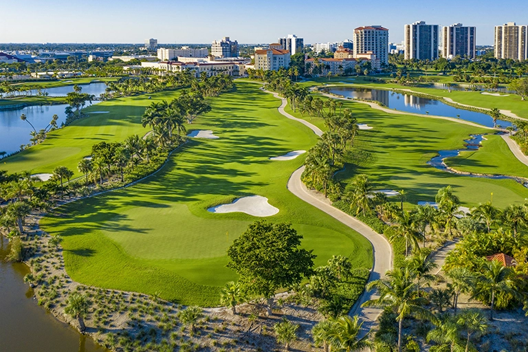 2026 News Golf 1 Aerial view of a coastal golf course with water, palm trees, and city buildings.