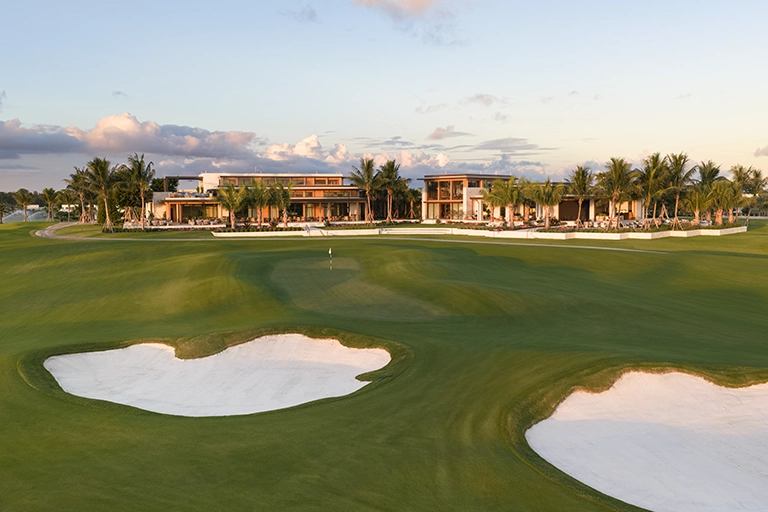 2026 News Golf 2 Golf course green and sand bunkers in front of a clubhouse at sunset.
