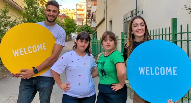 2026 Albania Lead Image Two pairs of Best Buddies participants and volunteers standing together outdoors, holding colorful ‘Welcome’ signs.