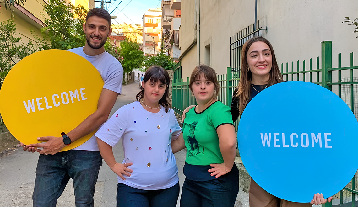 2026 Albania Lead Image Two pairs of Best Buddies participants and volunteers standing together outdoors, holding colorful ‘Welcome’ signs.